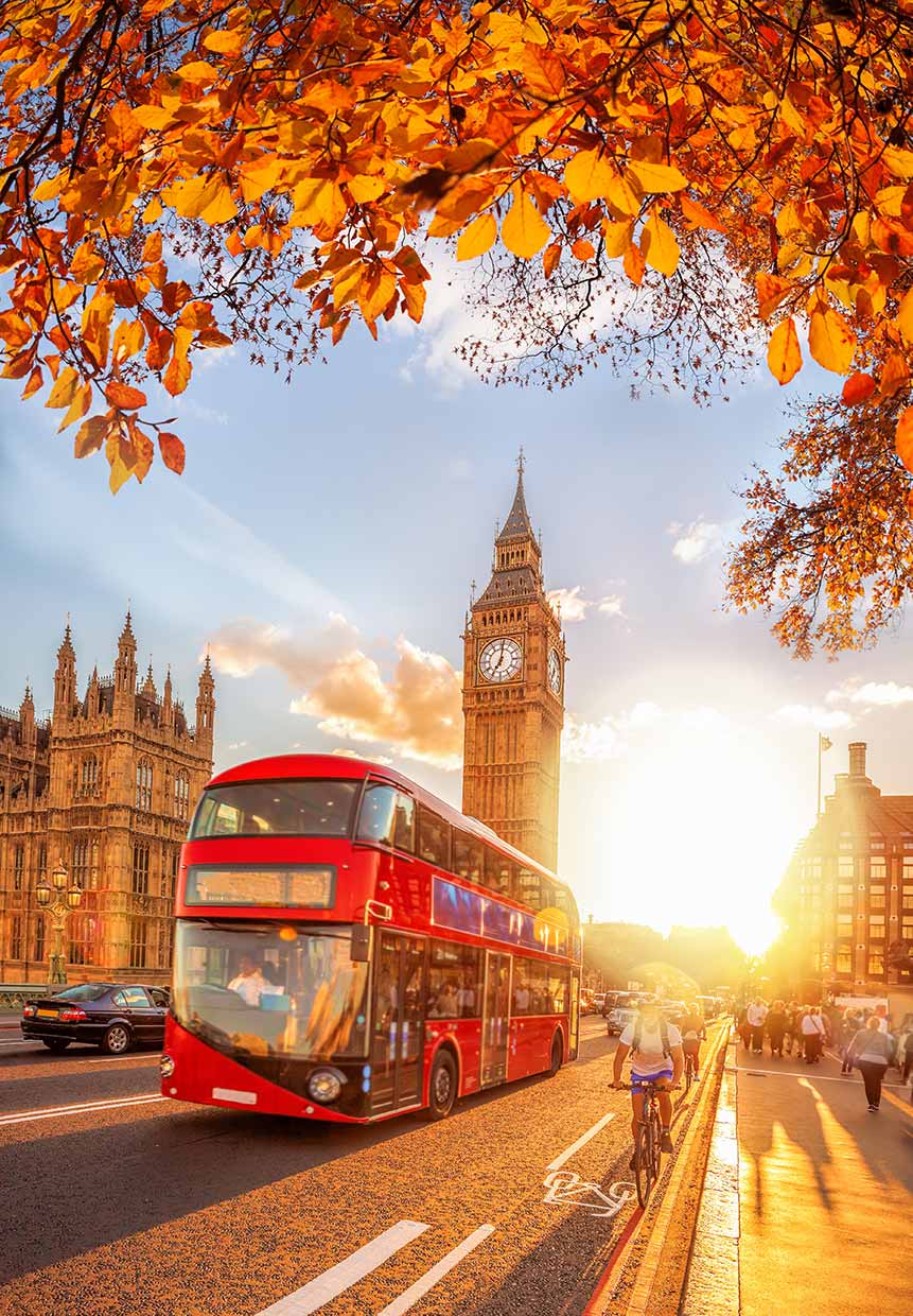 Red double-decker bus and autumn leaves with Big Ben and the Westminster area of London in the background during a sunset.