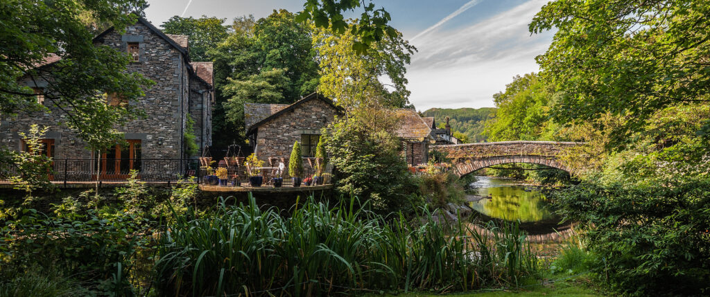 Scenic view of a historic stone bridge over a tranquil river at Grasmere, Lake District, England, surrounded by lush greenery and traditional stone buildings.