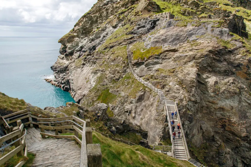 Wooden stairs winding down a rugged cliff at Tintagel Castle in Cornwall, UK, overlooking the turquoise waters of the Atlantic Ocean.