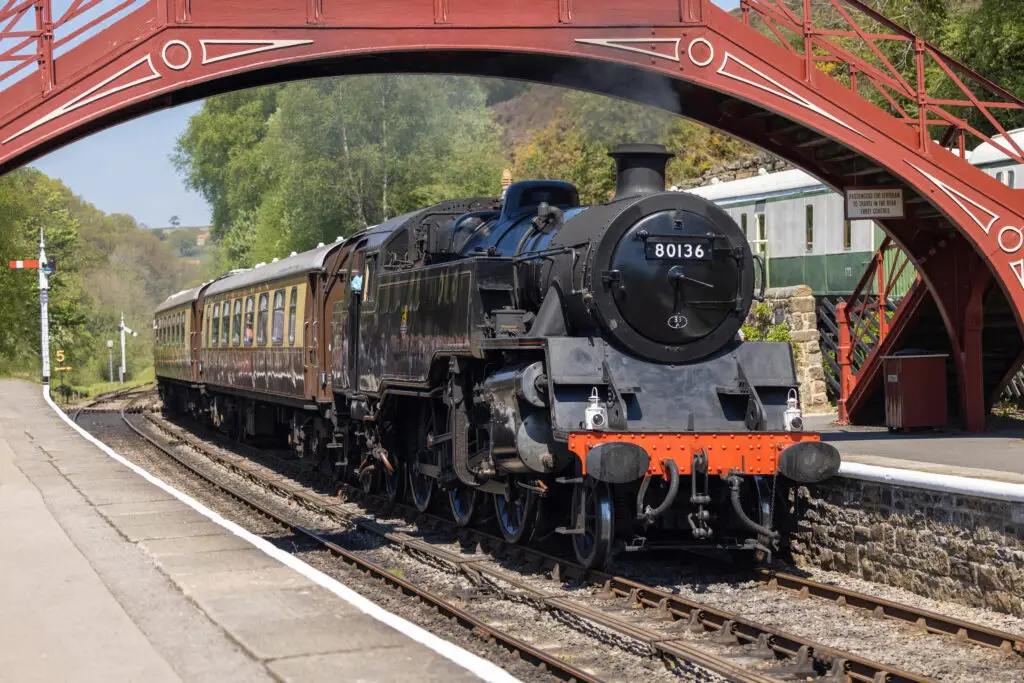Vintage steam train 80136 departing from Goathland Station in England under a historic red bridge, surrounded by lush greenery, ideal for enthusiasts of railway heritage tours.