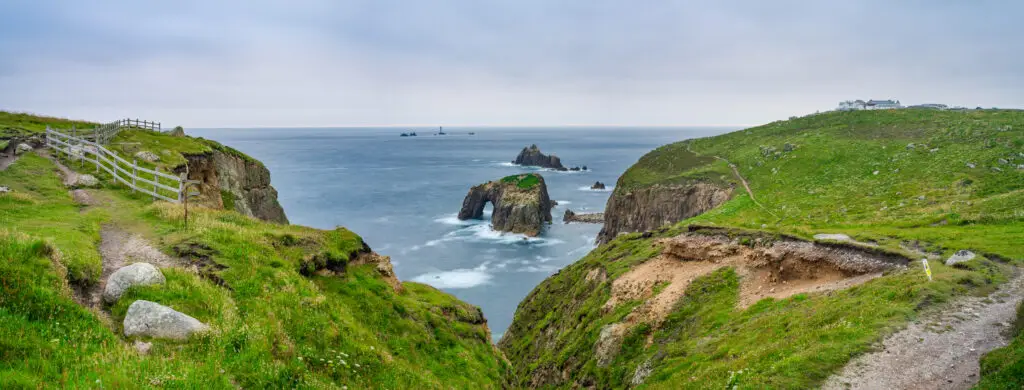 Panoramic view of Enys Dodman Arch near Land's End, Cornwall, showcasing rugged cliffs and a serene ocean under cloudy skies, a top destination for exploring natural British landscapes.