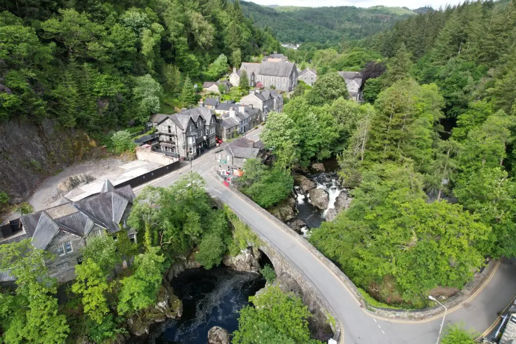 Aerial view of Betws-y-Coed, showing lush greenery, traditional stone buildings, and a river flowing through a scenic village in North Wales, UK.