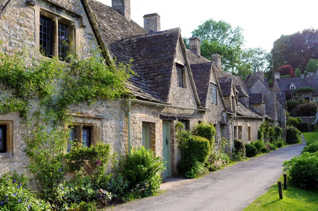 Scenic view of traditional old stone houses with slate roofs in the picturesque English countryside of the Cotswolds, featuring lush greenery and a quaint village road.